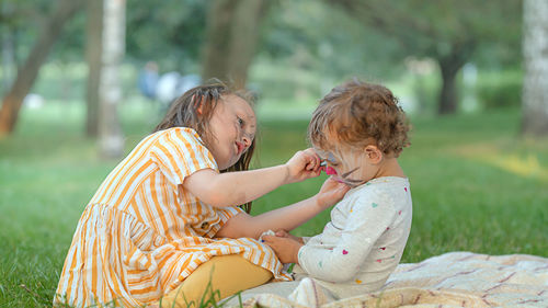 Girl painting on sisters face sitting at park