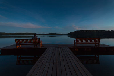 Rear view of man sitting on bench on pier over lake against blue sky at dusk