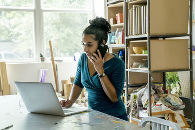 Businesswoman talking on smart phone while using laptop computer in office