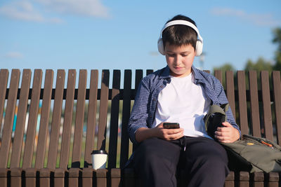 A cute boy teenager in headphones , watches mobile phone and sits on bench outdoor in the park 