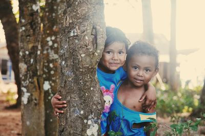 Portrait of siblings playing by tree trunk at park