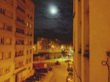 Illuminated street by buildings against sky at night