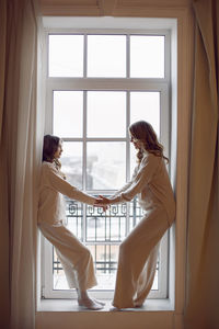 Family mother and daughter in white suit stand at the big window of the house on christmas in winter