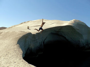 Rear view of man jumping on rock against clear blue sky