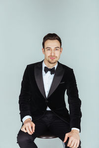 Portrait of confident young man in tuxedo sitting against blue background