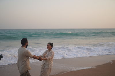 Rear view of couple standing at beach against sky during sunset