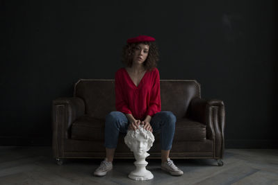 Portrait of woman with statue sitting on sofa against wall
