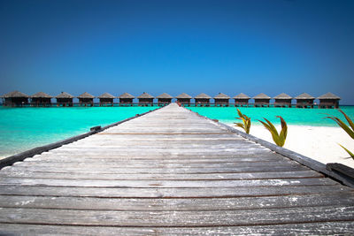 Jetty leading towards stilt houses amidst sea against clear blue sky on sunny day