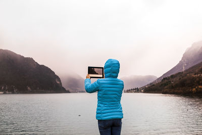 Rear view of woman photographing with digital tablet by lake during foggy weather
