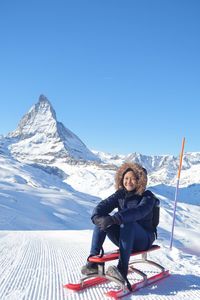 Full length of smiling teenage girl sitting on sled against snowcapped mountains during winter