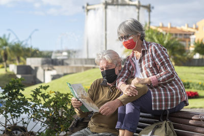 Senior couple wearing mask looking at map while sitting on bench outdoors