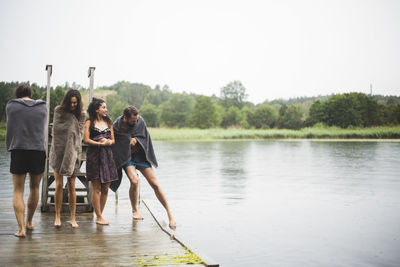 Male and female friends wrapped in towels walking on jetty over lake against clear sky