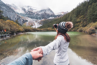 Cropped hand of man holding woman hand at lake against mountains