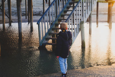 Boy standing on sea shore by pier during sunset