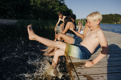 Happy shirtless boy splashing water while sitting with friends on jetty at summer camp