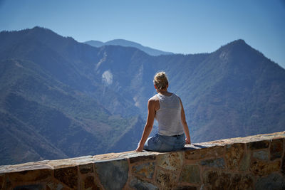 Rear view of woman sitting on retaining wall against mountain