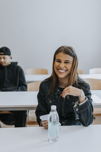 Portrait of smiling teenage girl sitting with smart phone and water bottle at desk in classroom