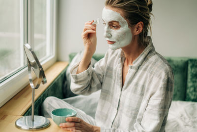 A woman in pajamas applies a clay mask to her face with a brush while looking in the mirror.