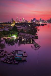 Illuminated buildings by river against sky at sunset