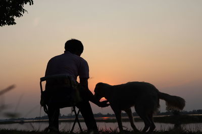 Rear view of man with dog standing against sky during sunset