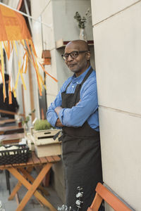 Portrait of male owner standing with arms crossed against wall at store entrance