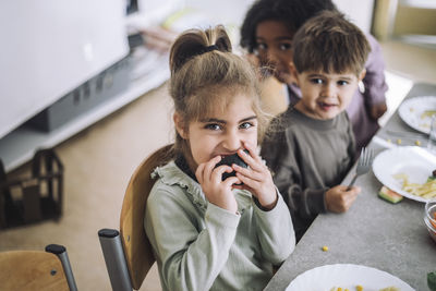 Portrait of girl biting fruit while sitting at dining table with mates in kindergarten