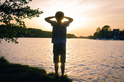 Rear view of boy standing by lake during sunset