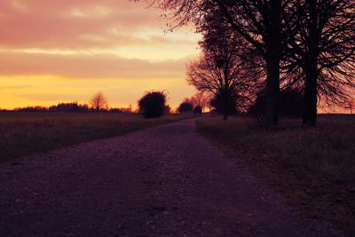 Dirt road amidst trees against sky during sunset