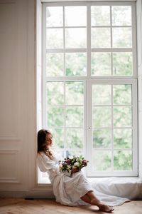 Young woman sitting with flowers on window at home