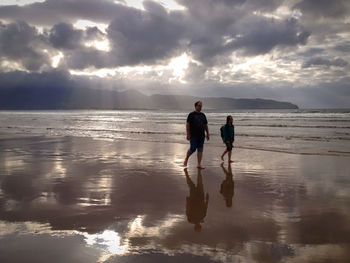 Full length of father with daughter walking at beach against cloudy sky