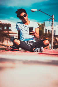 Young man using mobile phone while sitting against sky