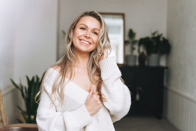 Blonde smiling woman face and hands with long hair in cozy knitted cardigan at the bright interior