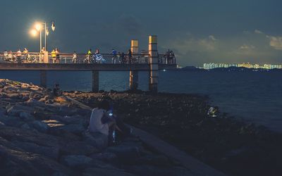 People sitting on rock next to jetty against cloudy sky