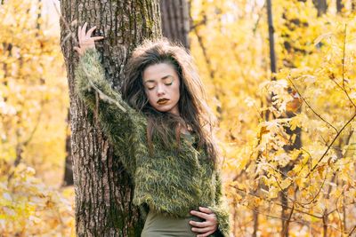Young woman standing against tree trunk in forest during autumn