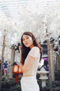 Portrait of beautiful young woman standing against white flowering trees at park