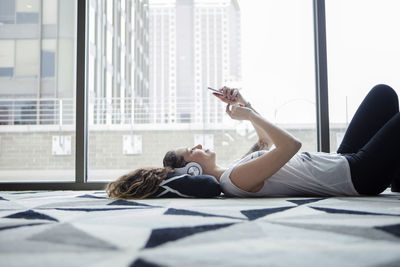 Young female using smart phone while lying on carpet by window at home