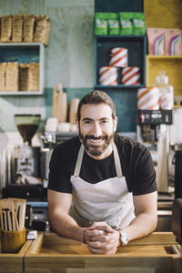 Portrait of smiling male retail clerk with hands clasped leaning on checkout counter at grocery store