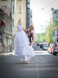 Portrait of young woman holding wedding dress while standing on street
