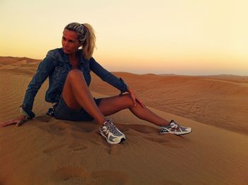 Woman sitting on sand while looking away at desert during sunset