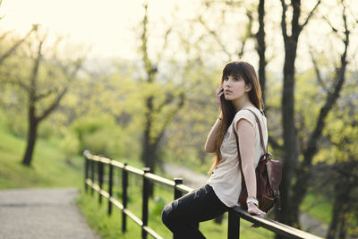 Woman looking away while sitting on railing against trees