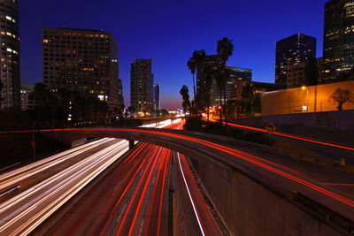 Light trails on road by illuminated buildings against sky at night