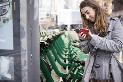 Young woman holding credit card while using smart phone by bike share at parking lot