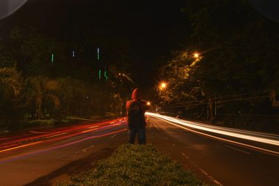 Rear view of man standing amidst light trails on road at night