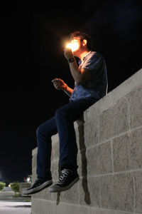 Side view of man smoking while sitting on wall at night
