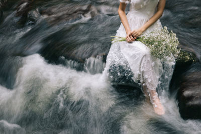 Low section of woman in wedding dress sitting on rock over river