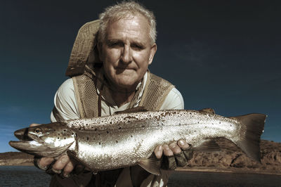 Portrait of senior man holding fish while standing against blue sky