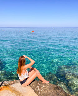 Rear view of woman sitting on rock by sea against clear sky