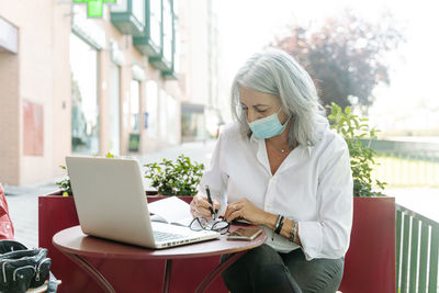 Busy mature female entrepreneur in mask sitting in outdoors cafe and taking notes while working remotely in city during coronavirus pandemic