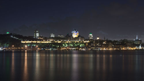 Illuminated buildings by river against sky at night