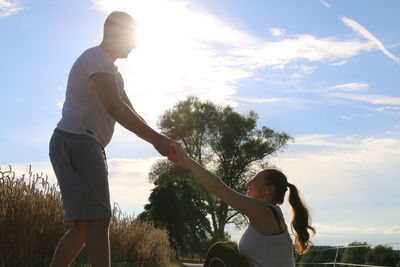 Side view of man holding woman hands against tree and sky